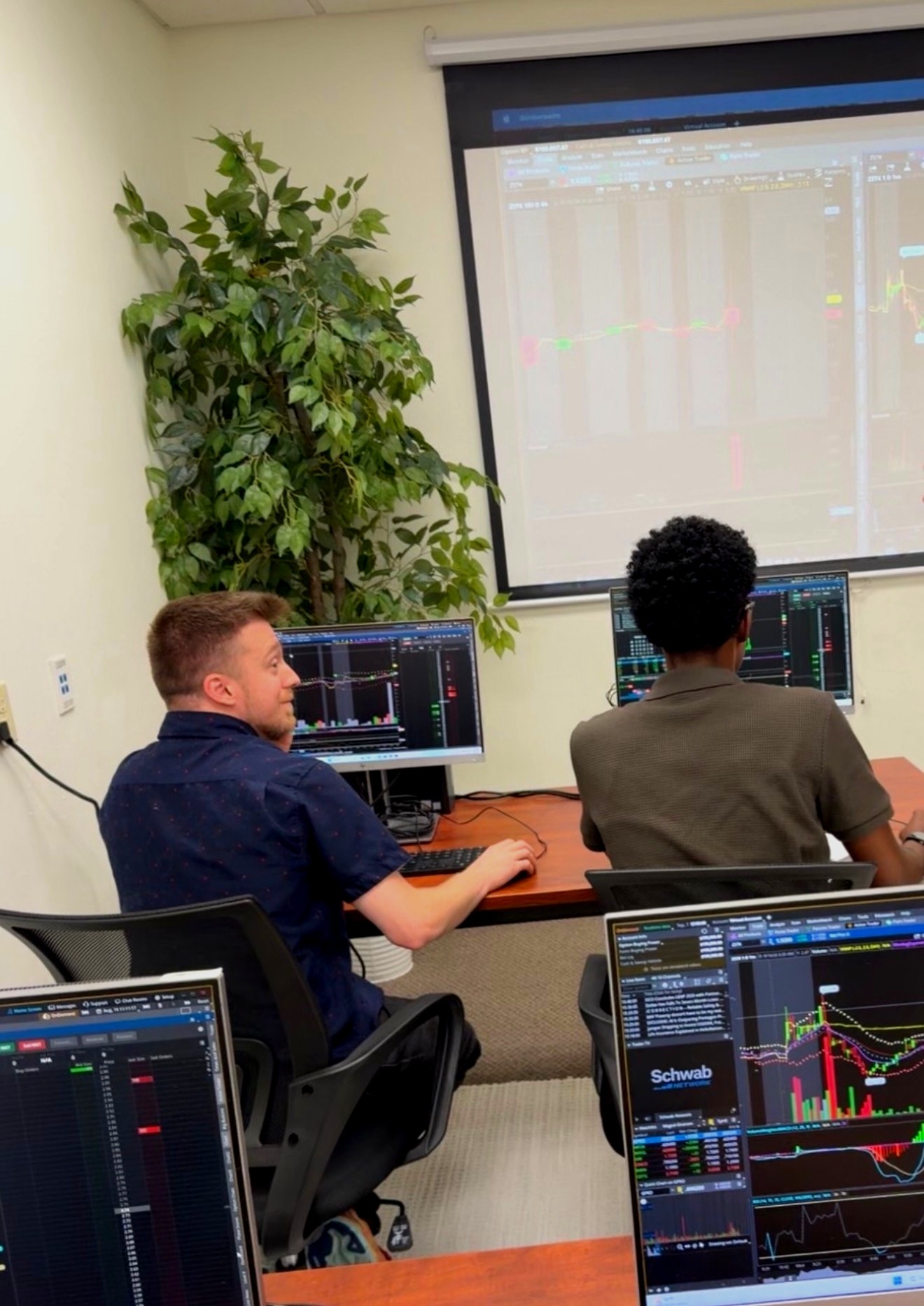 Two beginner students at their Schwab trading stations at MAAL TRADING ACADEMY, a live chart on the big screen behind them.
