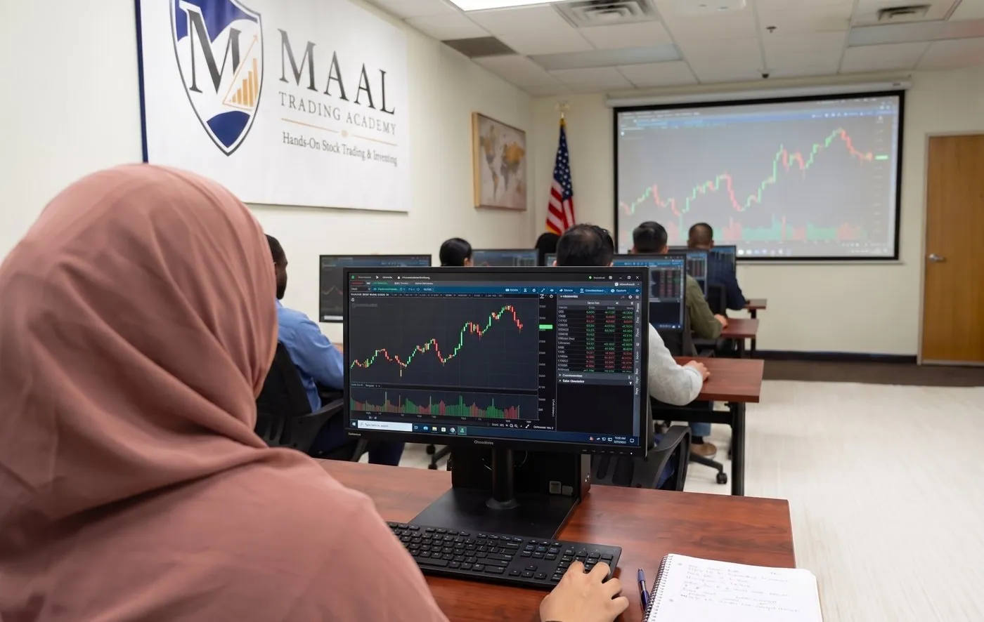A student at a trading station, a price chart and an order ticket on screen, notes open beside the keyboard.